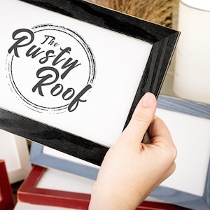 May include: A black framed picture with the text "The Rusty Roof" in a circular design. Other frames in red and blue are visible in the background. The frame is being held by a hand.