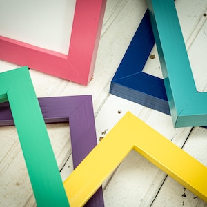 May include: A collection of colorful picture frames in various shades, including pink, green, purple, yellow, blue, and teal. The frames are arranged on a white wooden surface, showcasing their vibrant colors and geometric shapes.
