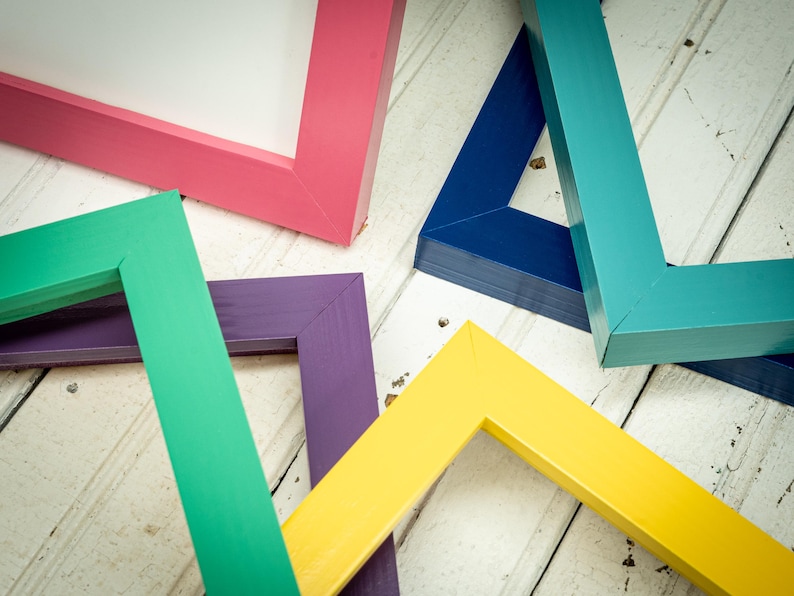 May include: A collection of colorful picture frames in various shades, including pink, green, purple, yellow, blue, and teal. The frames are arranged on a white wooden surface, showcasing their vibrant colors and geometric shapes.