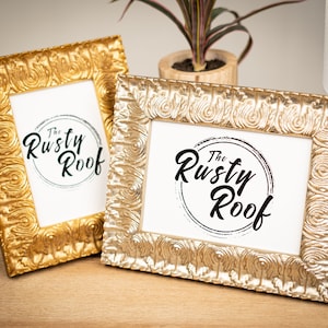 May include: Two gold-colored picture frames with ornate designs. The frames are on a wooden surface. The frames contain a white background with black text that reads "The Rusty Roof".