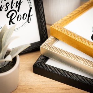 May include: Three picture frames with rope-like detailing around the edges. The frames are stacked on top of each other, with the black frame on the bottom, the silver frame in the middle, and the gold frame on top. The top frame has the text "The Rusty Roof" printed on the inside.