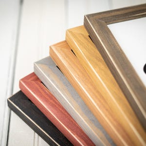 May include: A stack of framed art pieces in various colors, including black, red, gray, light brown, and dark brown. The frames are made of wood and have a simple, modern design. The artwork inside is partially visible.