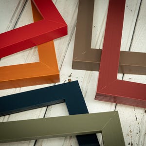 May include: An assortment of colorful picture frames in various shades, including red, orange, navy blue, olive green, and brown. The frames are arranged on a light-colored wooden surface, showcasing their angular design and glossy finish.