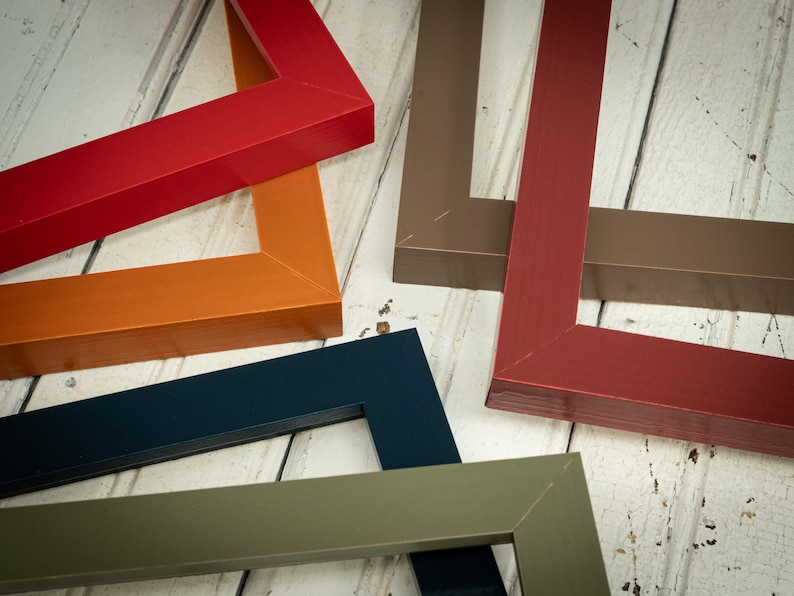 May include: An assortment of colorful picture frames in various shades, including red, orange, navy blue, olive green, and brown. The frames are arranged on a light-colored wooden surface, showcasing their angular design and glossy finish.
