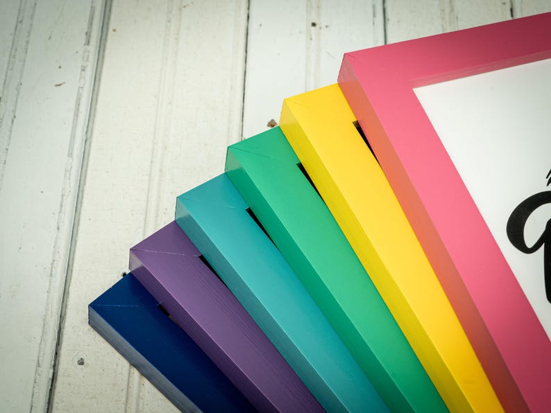 May include: A collection of colorful picture frames in a rainbow of hues, including pink, yellow, green, and blue. The frames are arranged diagonally, showcasing their vibrant colors against a white wooden background. One frame displays a portion of black script.