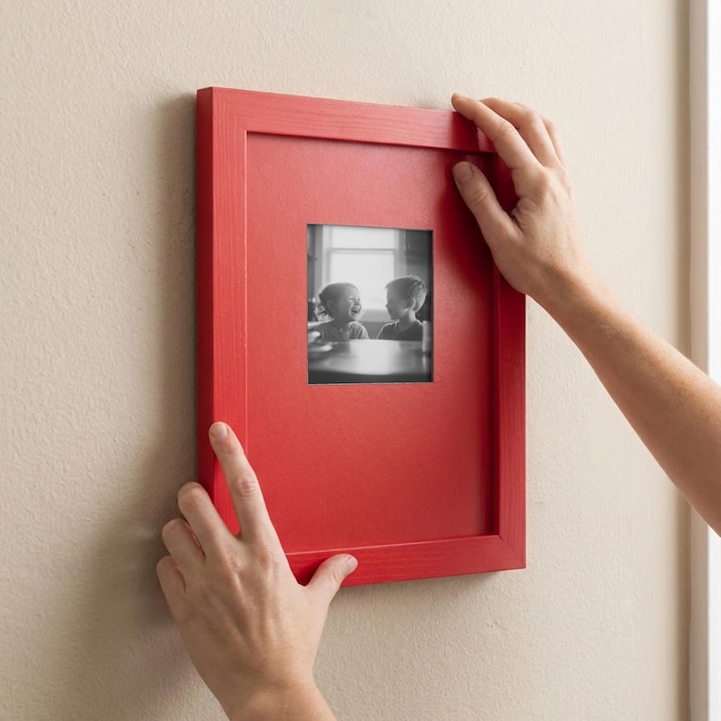 A person's hands are holding a red picture frame with a black and white photograph of two children inside it, hanging on a wall.