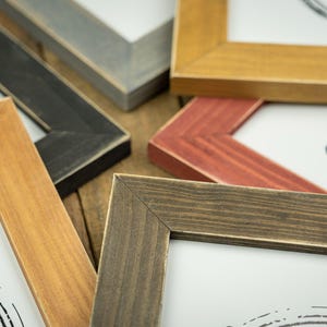May include: Close-up of several wooden picture frames in various colors, including black, brown, red, and gold. The frames are arranged at different angles, showcasing their corners and the white artwork within.