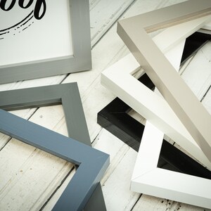 May include: A collection of various picture frames in shades of gray, white, and black, arranged on a white wooden surface. One frame displays the text "Roof" in a black, stylized font. The frames are of different sizes and shapes.