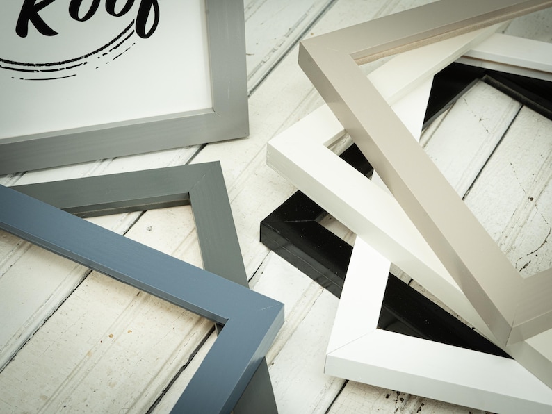 May include: A collection of various picture frames in shades of gray, white, and black, arranged on a white wooden surface. One frame displays the text "Roof" in a black, stylized font. The frames are of different sizes and shapes.