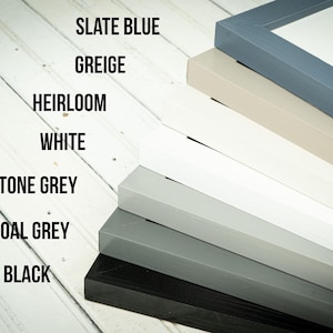May include: A stack of painted wooden picture frames in various colors, including slate blue, greige, heirloom, white, stone grey, charcoal grey, and black. The frames are angled, showcasing the different color options.