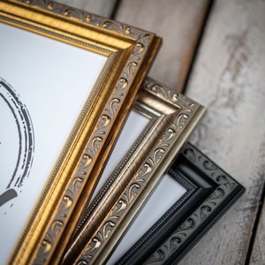May include: Three picture frames stacked on top of each other. The top frame is gold with an ornate design. The middle frame is silver with an ornate design. The bottom frame is black with a simple design.