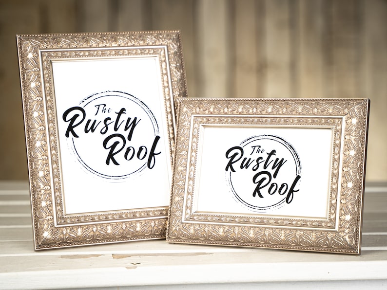 May include: Two ornate gold picture frames with a white background and the text "The Rusty Roof" in black script. The frames are on a white wooden surface.