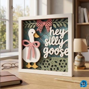 Puede incluir: Una obra de arte enmarcada con un marco blanco. La obra presenta un ganso blanco con un lazo rosa, un lazo rosa en la parte superior y las palabras "hey silly goose" en cursiva. El fondo tiene un patrón de azulejos turquesa y recortes florales.