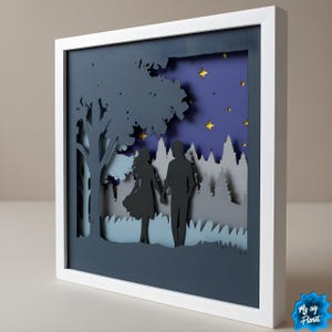 Könnte beinhalten: Ein gerahmtes Papierkunstwerk, das ein Paar zeigt, das sich unter einem Baum an den Händen hält, mit einem Wald und einem Sternenhimmel im Hintergrund. Das Kunstwerk verwendet geschichtetes Papier in Blau-, Grau- und Goldtönen, eingebettet in einen weißen Rahmen.