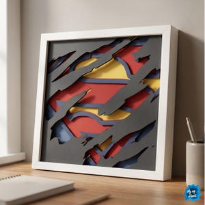 May include: A framed 3D paper art piece featuring the Superman logo. The artwork has a white frame and a layered design with red, yellow, and blue colors. The design gives the illusion of a ripped shirt revealing the iconic symbol.