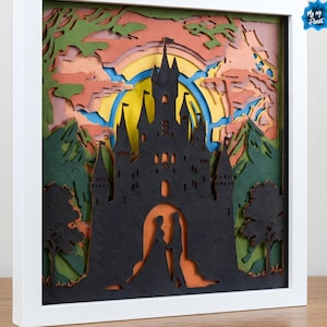 Puede incluir: Una obra de arte en papel multicapa enmarcada que presenta una silueta de castillo negro contra una colorida puesta de sol. La obra de arte incluye un sol amarillo, cielo naranja, árboles verdes y una pareja en el arco del castillo. El marco es blanco.