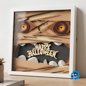 May include: A square white-framed shadow box featuring a layered paper art design of a mummy face with yellow eyes and sharp white teeth. The center of the open mouth displays the text Happy Halloween in a stylized beige font against a dark background.
