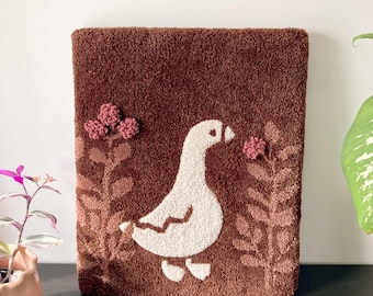 May include: A brown plush wall hanging featuring a white goose and pink flowers. The goose is facing right and has a long neck and beak. The flowers are in the top left and right corners of the hanging.