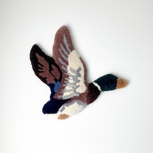 May include: A handcrafted wall hanging depicting a duck in flight. The artwork uses a palette of brown, blue, and white, with a green head and tan beak. The wings are outstretched, and the piece is set against a white backdrop.
