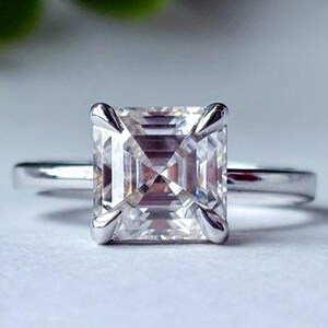 May include: A silver ring featuring a square-cut diamond. The diamond is held in place by four prongs. The ring is presented on a white surface, with a blurred green background of foliage.