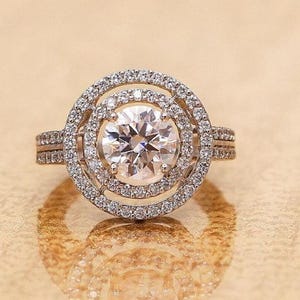 May include: A diamond engagement ring with a large, round center stone, surrounded by two halos of smaller diamonds. The ring is set on a band with additional diamond accents. The ring is displayed on a reflective surface.
