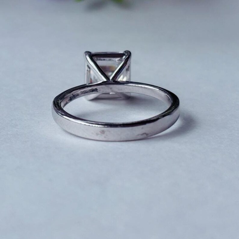 May include: A silver ring featuring a large, square-cut clear gemstone. The ring's band is smooth and polished, and the gemstone is held in place by four prongs. The ring is set against a soft, light background.