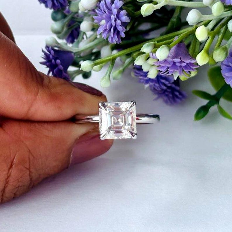 May include: A silver ring featuring a square-cut diamond. The diamond is clear and set with four prongs. The ring is shown against a backdrop of purple and white flowers, with a focus on the ring.