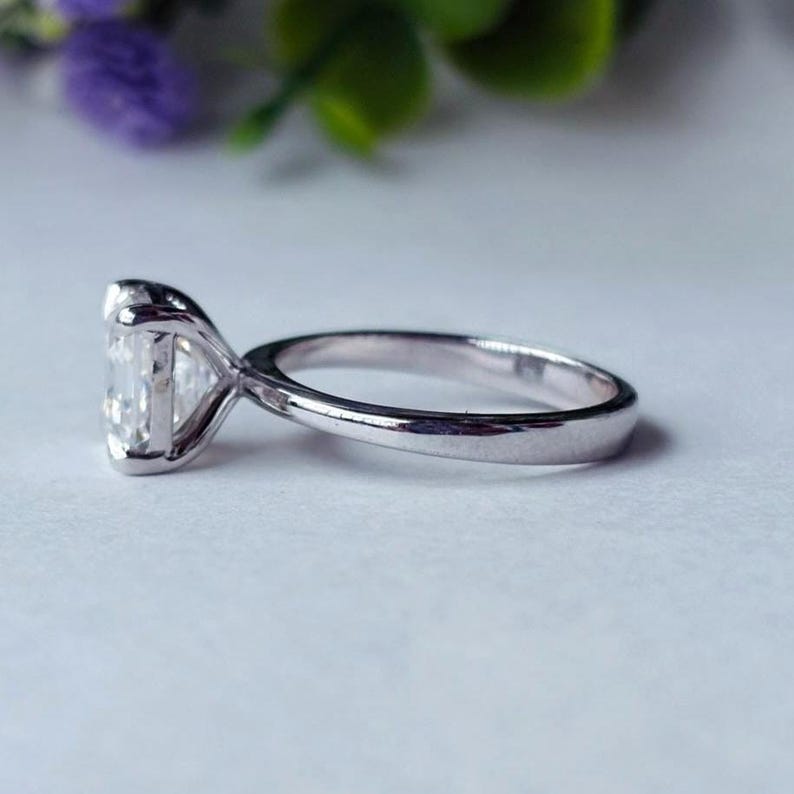 May include: A silver ring featuring a large, rectangular, clear gemstone. The ring has a classic four-prong setting, and the band is smooth and polished. The side view showcases the gemstone's facets and the ring's elegant design.