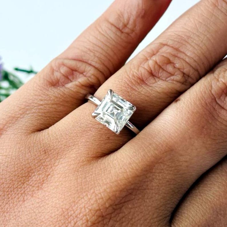 May include: A silver ring featuring a square-cut, clear gemstone. The gemstone is held in place by four prongs. The ring is displayed on a hand.
