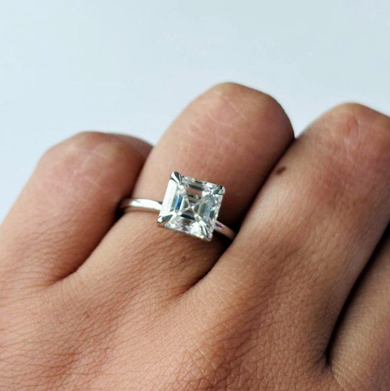 May include: A silver ring featuring a square-cut diamond. The clear gemstone is the main feature, reflecting light. The ring is displayed on a person's finger, against a plain background. A classic piece of jewellery.
