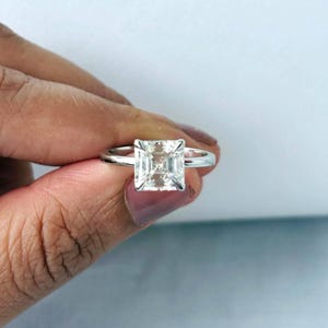 May include: A silver ring featuring a large, square-cut, clear gemstone. The gemstone is held in place by a four-prong setting, with a thin, polished band. The ring is displayed against a white and blue backdrop.