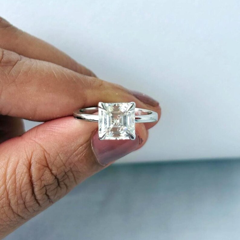 May include: A silver ring featuring a large, square-cut, clear gemstone. The gemstone is held in place by a four-prong setting, with a thin, polished band. The ring is displayed against a white and blue backdrop.