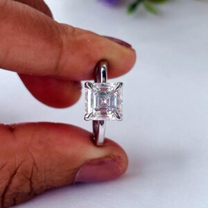 May include: A silver ring featuring a large, square-cut, clear gemstone. The gemstone is secured by four prongs. The ring is held between two fingers against a blurred background.