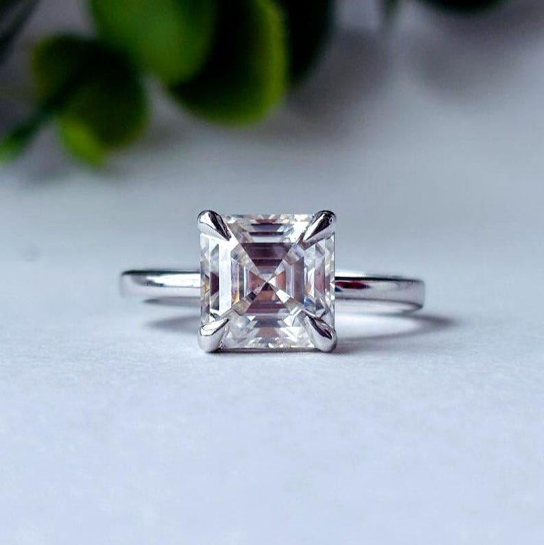 May include: A silver ring featuring a square-cut diamond. The diamond is held in place by four prongs. The ring is presented on a white surface, with a blurred green background of foliage.
