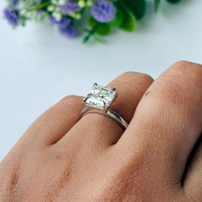 May include: A silver ring featuring a large, square-cut, clear gemstone. The ring is on a finger, with a blurred background of purple flowers and green leaves.