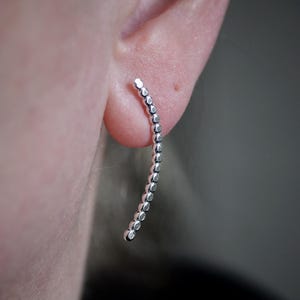 May include: A silver ear climber earring with a curved design. The earring is adorned with a series of small, round, silver beads. The earring is shown on an ear, with a blurred background.