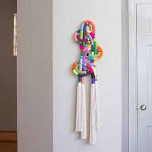 May include: A colorful wall hanging with a knotted rope design. The artwork features a variety of bright colors, including pink, orange, green, and blue. Three white tassels hang from the bottom of the piece. This decorative item is a unique piece of wall art.