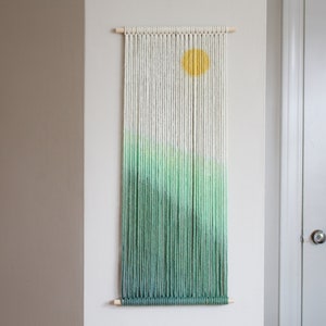 May include: A green and white macrame wall hanging with a yellow circle in the center. The hanging is made of yarn and is attached to a wooden dowel.