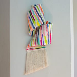 May include: A woven wall hanging featuring a vibrant, multi-colored striped pattern in shades of neon pink, bright yellow, orange, light blue, and dark blue. The textile piece has a textured surface and ends in a long, cream-colored fringe.