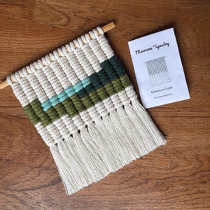 May include: A macrame tapestry featuring off-white, olive green, and light turquoise knotted cord patterns suspended from a wooden dowel, accompanied by a white paper booklet titled Macrame Tapestry Reference Guide by Sonya Benham.