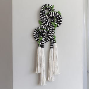 May include: Black and white macrame wall hanging with green leaves and white tassels.