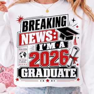 May include: White sweatshirt with a graduation-themed graphic. The design features the text "BREAKING NEWS: I'M A 2026 GRADUATE" in black and red, along with a graduation cap and diploma. The sweatshirt has a crew neck.