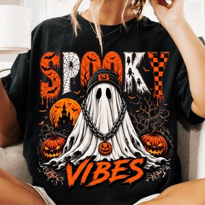 May include: A black t-shirt featuring a graphic of a ghost wearing a beanie and a chain necklace. The design includes the text "SPOOKY VIBES" in orange, along with illustrations of a haunted house, pumpkins, bats, and a spider web in orange and white.