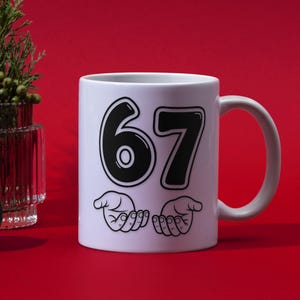 Pu&ograve; includere: Tazza in ceramica bianca con il numero "67" in nero, bordato di bianco. Sotto i numeri ci sono due mani con i palmi aperti. La tazza ha un manico bianco ed &egrave; posta su uno sfondo rosso.