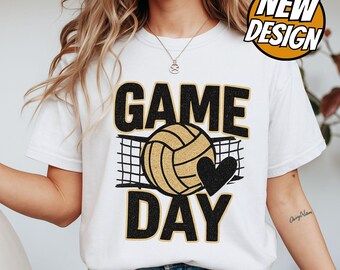 May include: White t-shirt with a volleyball-themed design. The shirt features the words "GAME DAY" in black and gold lettering, a volleyball, a net, and a black heart. A "NEW DESIGN" badge is in the upper right corner.