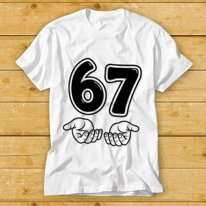 Pu&ograve; includere: T-shirt bianca con il numero "67" in grassetto nero con un contorno bianco. Sotto i numeri ci sono due mani stilizzate con i palmi aperti, anch'essi neri. La maglietta &egrave; realizzata in un materiale morbido.