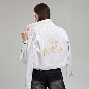 May include: White denim jacket with pearl embellishments on the shoulders. The back of the jacket features gold embroidered text that reads "Mrs. Miller" and the date "5/17/2022". The jacket has a classic button-up design.