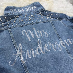 May include: Light blue denim jacket with pearl and rhinestone embellishments. The back of the jacket features the text "Mrs. Anderson" in rhinestones and the date "25.04.2026" in white.
