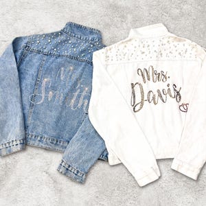 Op de afbeelding: Er worden twee spijkerjacks getoond, een lichtblauwe en een witte. De lichtblauwe jas heeft de naam "Mrs. Smith" in zilveren letters en de witte jas heeft "Mrs. Davis" in gouden letters. Beide jassen zijn versierd met parels.