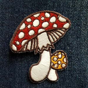 May include: Embroidered mushroom patch featuring a red cap with white spots, a white stem, and a smaller yellow mushroom. The patch has a brown outline and is set against a blue denim background. This patch is perfect for adding to clothing or accessories.
